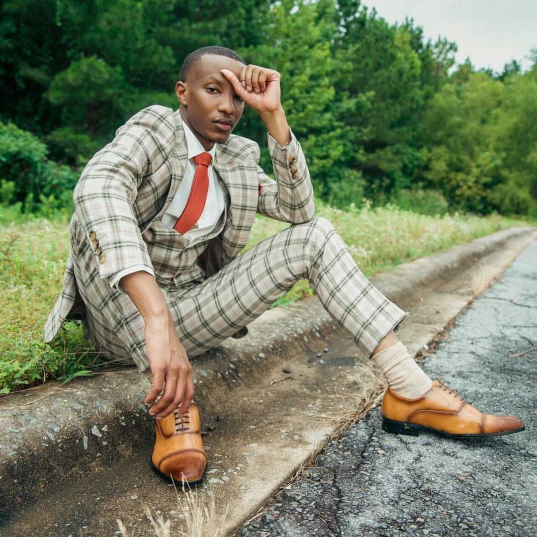 Man sitting outdoors in a light tan plaid 3-piece suit with a white dress shirt, red necktie, and cognac brown leather dress shoes by Dolce Vesta.