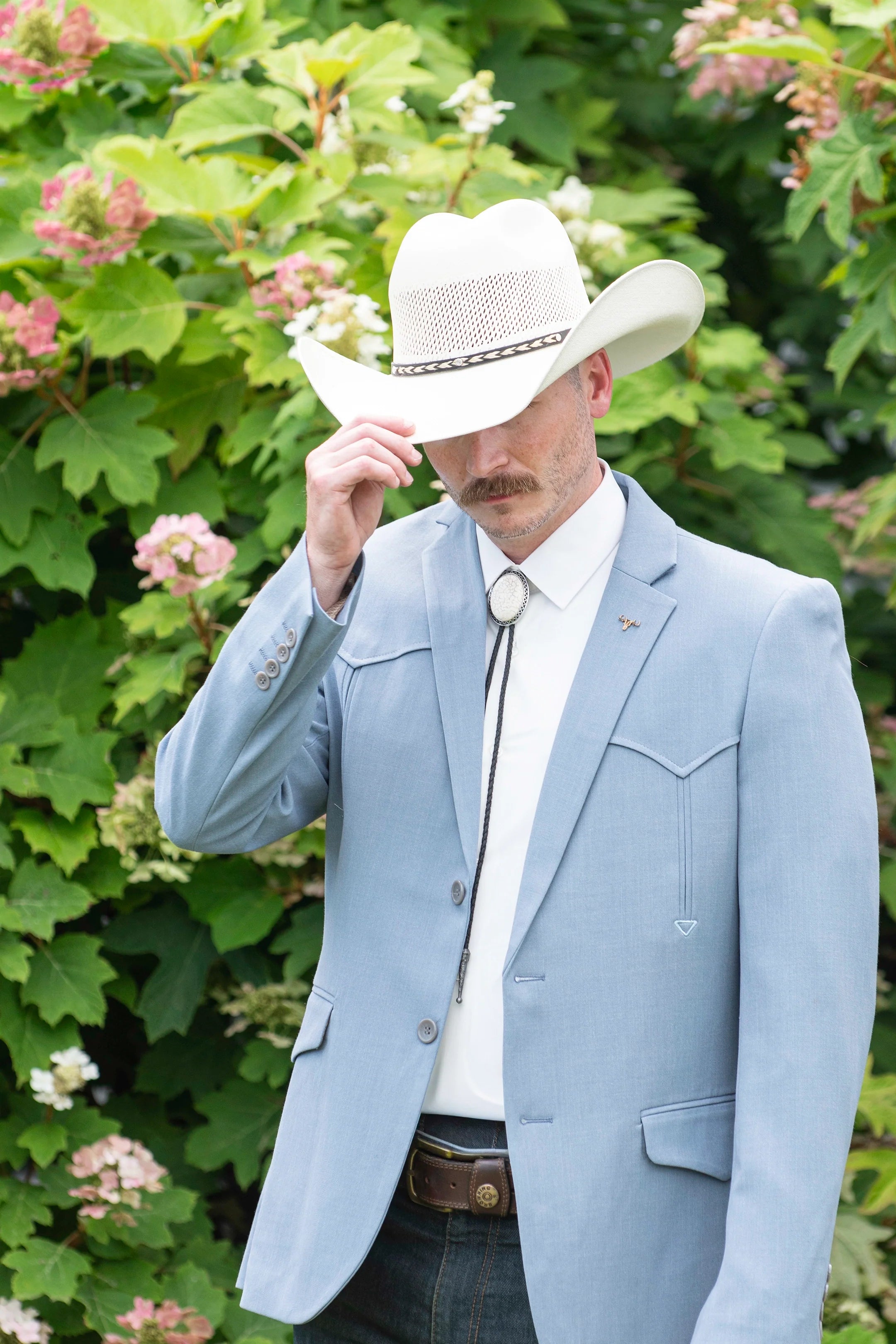 A man wearing a light blue Western suit jacket, white shirt, decorative bolo tie, and white cowboy hat tipping his hat down while standing outdoors.