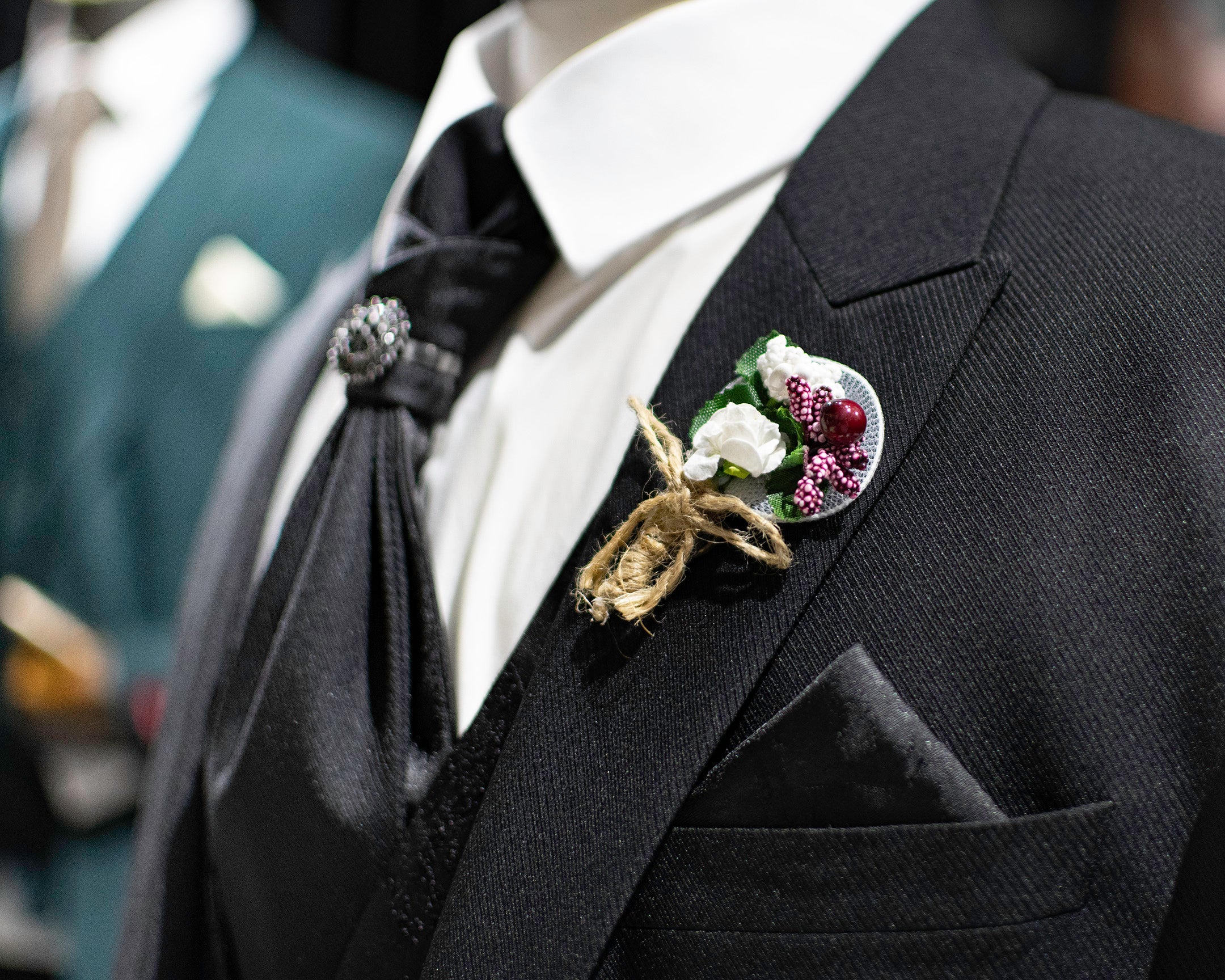 Black Suit with peak lapel with a bouquet Lapel Pin, black pocket square, and jeweled black tie on a mannequin from Dolce Vesta.