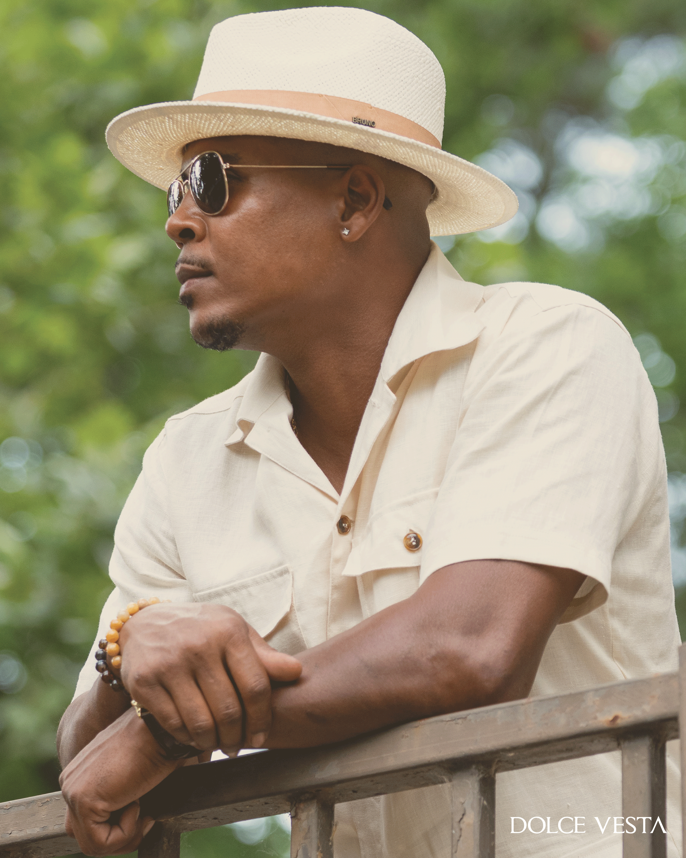 A man wearing a cream fedora hat, aviator sunglasses, a short-sleeve button-up shirt, and bracelets leaning on a railing outdoors.