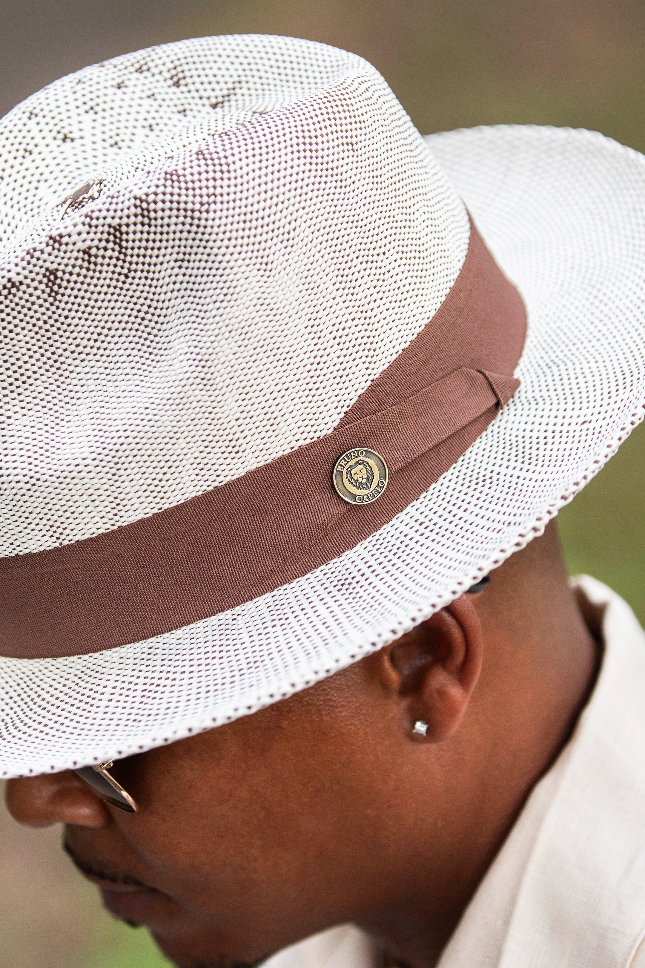 Close-up view of a man wearing a white straw Bruno Capelo fedora with a brown hatband and signature lion crest pin, available at Dolce Vesta.
