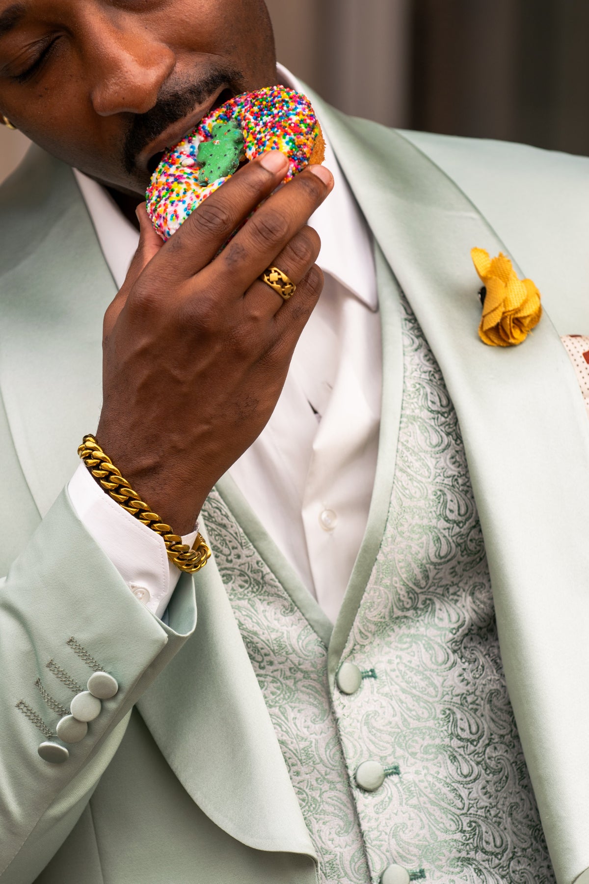 Close-up of a man in a mint green suit and paisley vest eating a rainbow sprinkle donut, wearing gold jewelry and a yellow lapel flower.