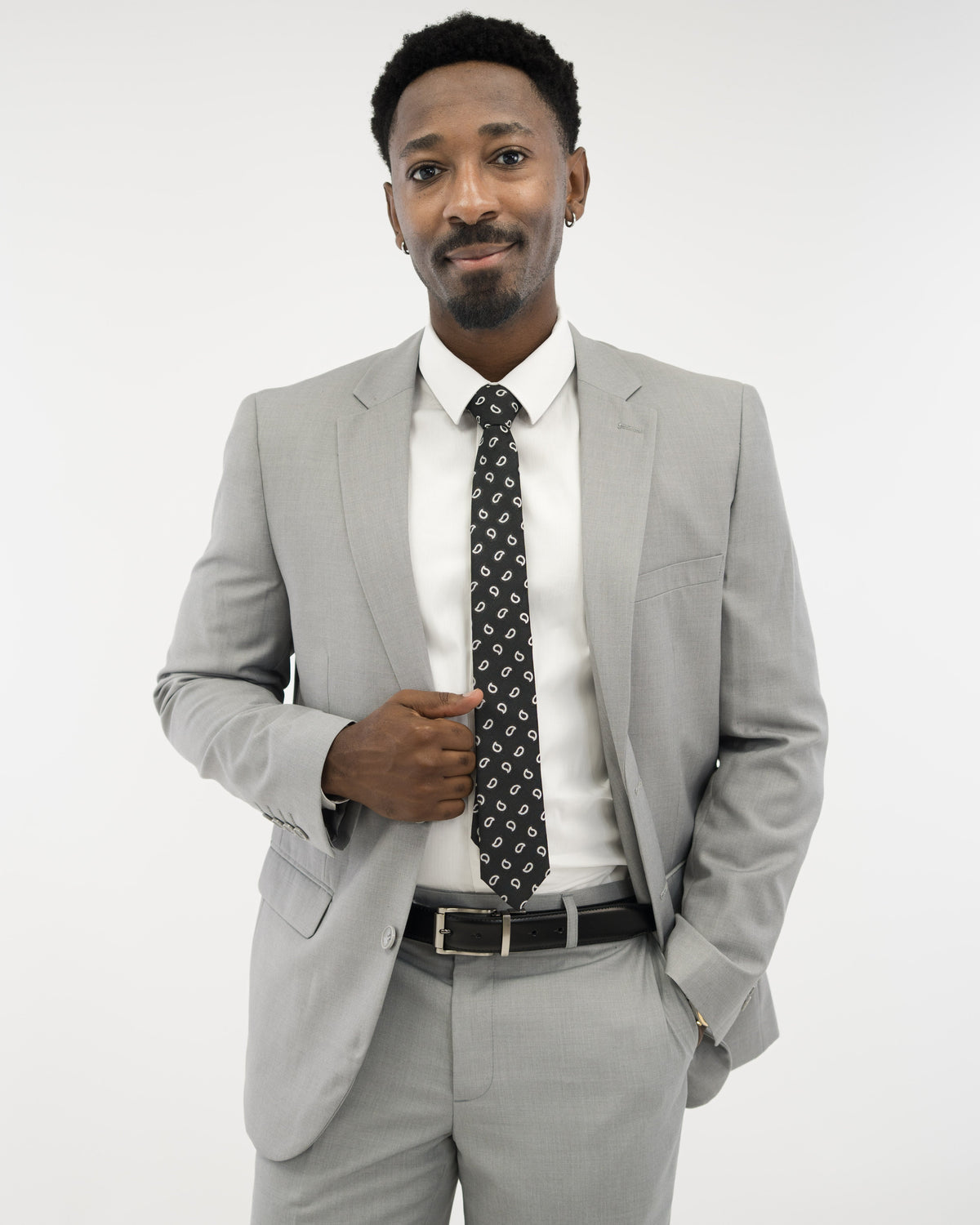 A Black man in a light gray suit, crisp white shirt, and black patterned tie, smiling against a plain white background.